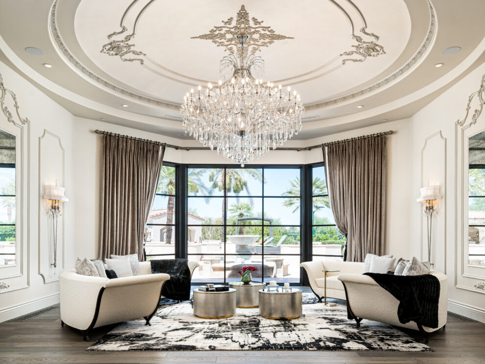 Living Room with a Chandelier and Bay Window: A luxurious living room featuring a grand chandelier hanging from an intricately designed ceiling. The room has elegant beige sofas, round metallic coffee tables, and floor-to-ceiling windows with a view of palm trees outside. The decor includes ornate wall moldings and a patterned area rug.